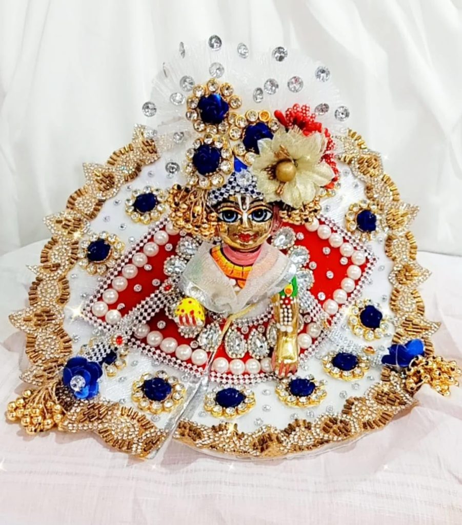 Laddu Gopal idol dressed in red and white designer poshak decorated with pearls, golden embroidery, blue floral accents, and jewel crown